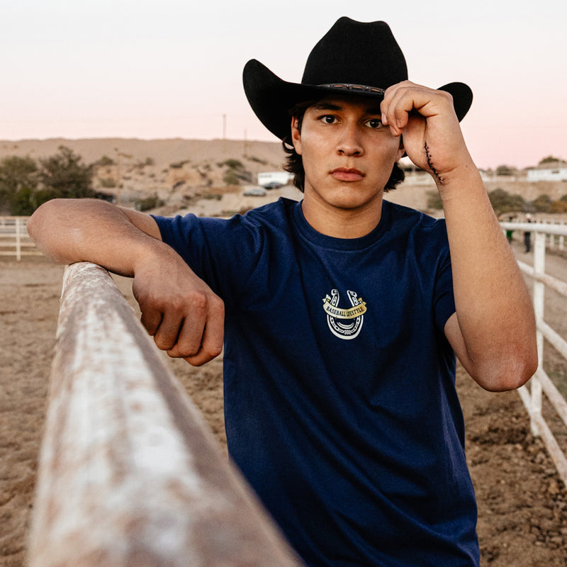 Person wearing a cowboy hat and blue shirt with a Baseball Lifestyle horseshoe logo, standing near a fence in an outdoor setting.