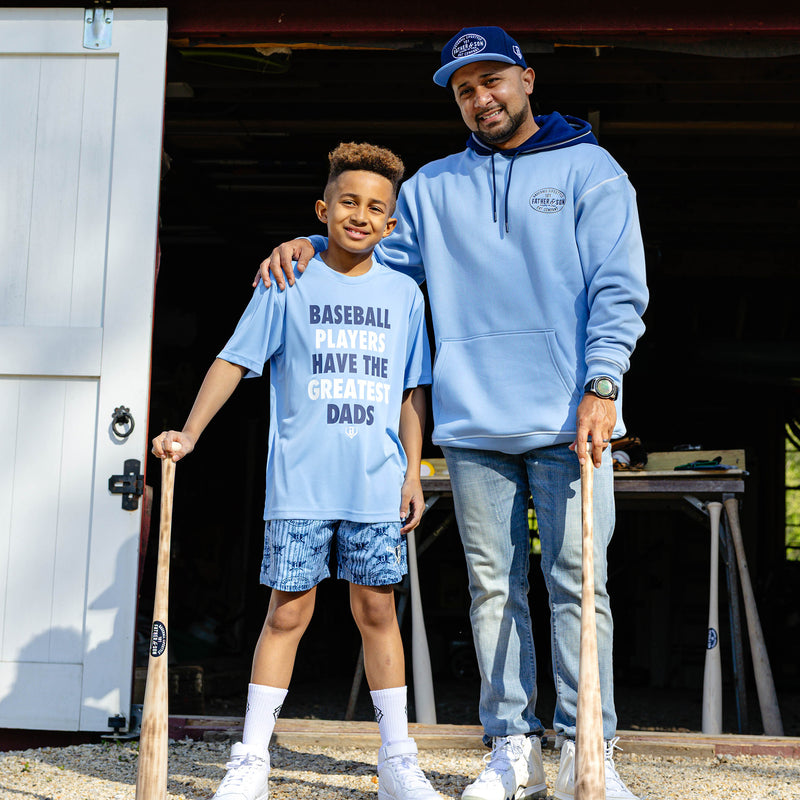 Father and son standing together outside a barn holding wooden baseball bats wearing Father & Son Bat Company collection pieces including the navy snapback and columbia blue hoodie.