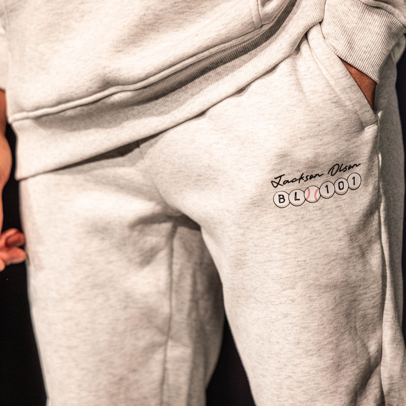 Close-up of the thigh graphic on the heather gray BSBL Era sweatpants, showing the "Jackson Olson" script above the BL101 baseball bead logo