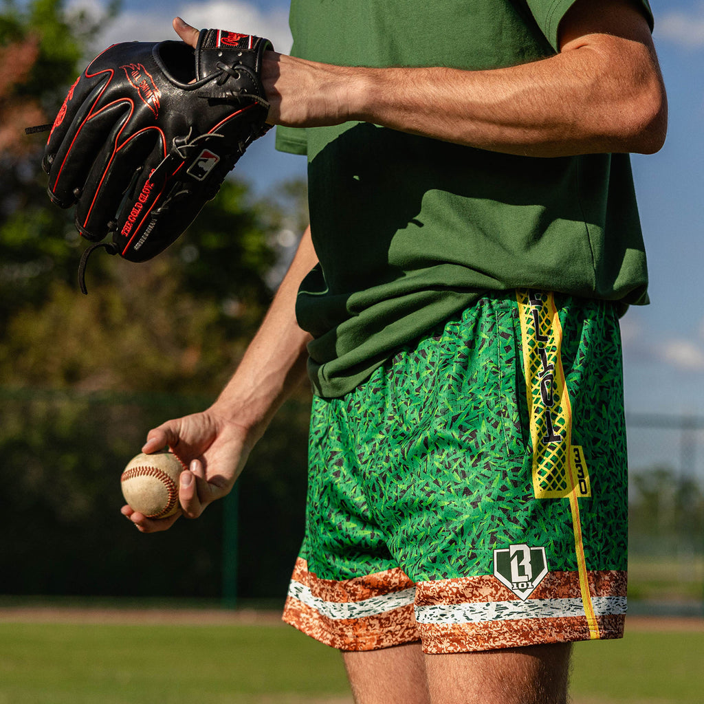 Close up of man holding a baseball and glove wearing green grass print mesh shorts with BL101 logo patch yellow foul pole side stripe and brown marbled hem
