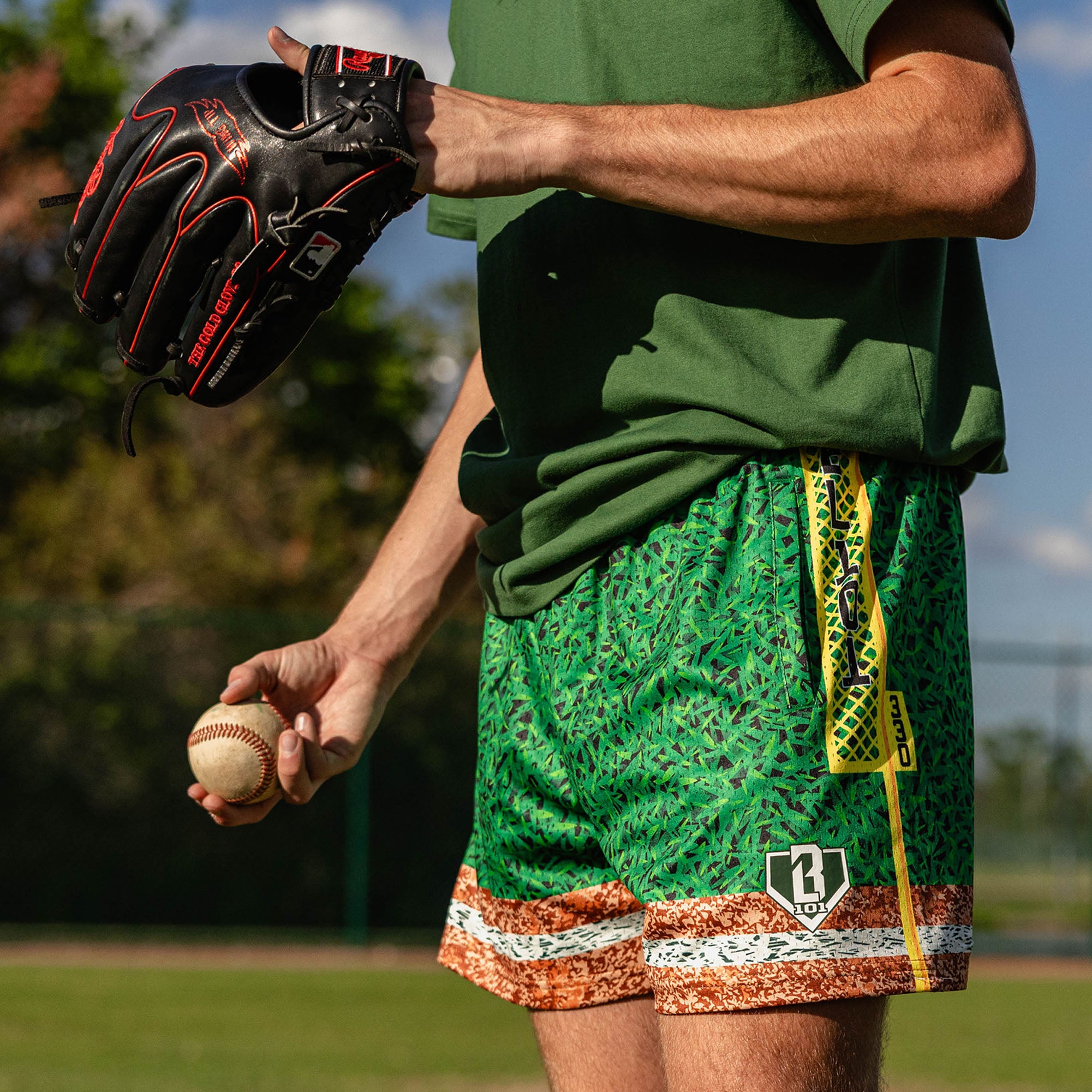 Close up of man holding a baseball and glove wearing green grass print mesh shorts with BL101 logo patch yellow foul pole side stripe and brown marbled hem
