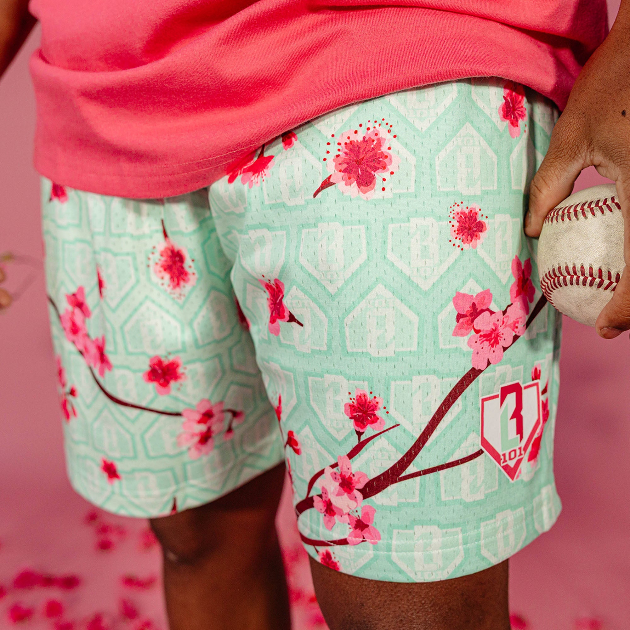 Close up of pink cherry blossom print and BL101 shield logo on mint green mesh shorts with home plate background pattern, worn by a male model holding a baseball