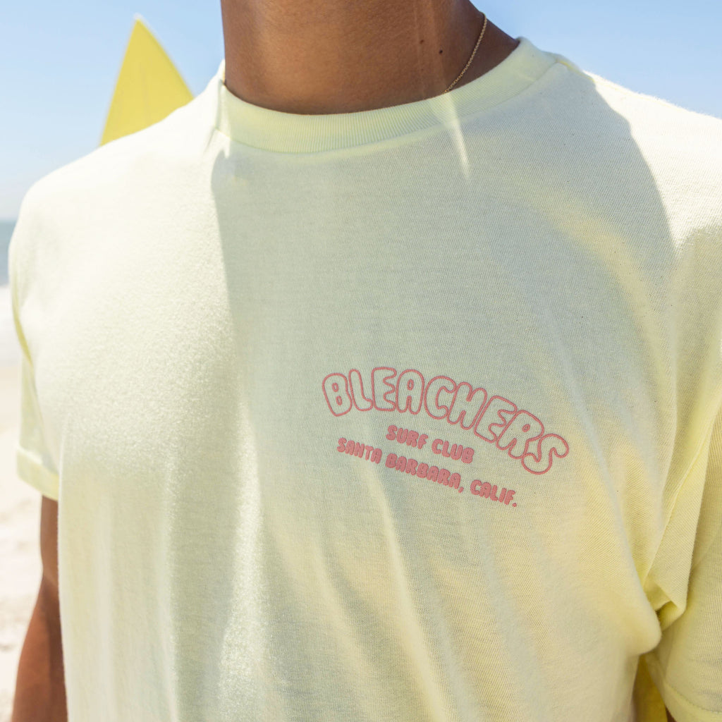 Adult model wearing yellow graphic tee with "Bleachers Surf Club" design