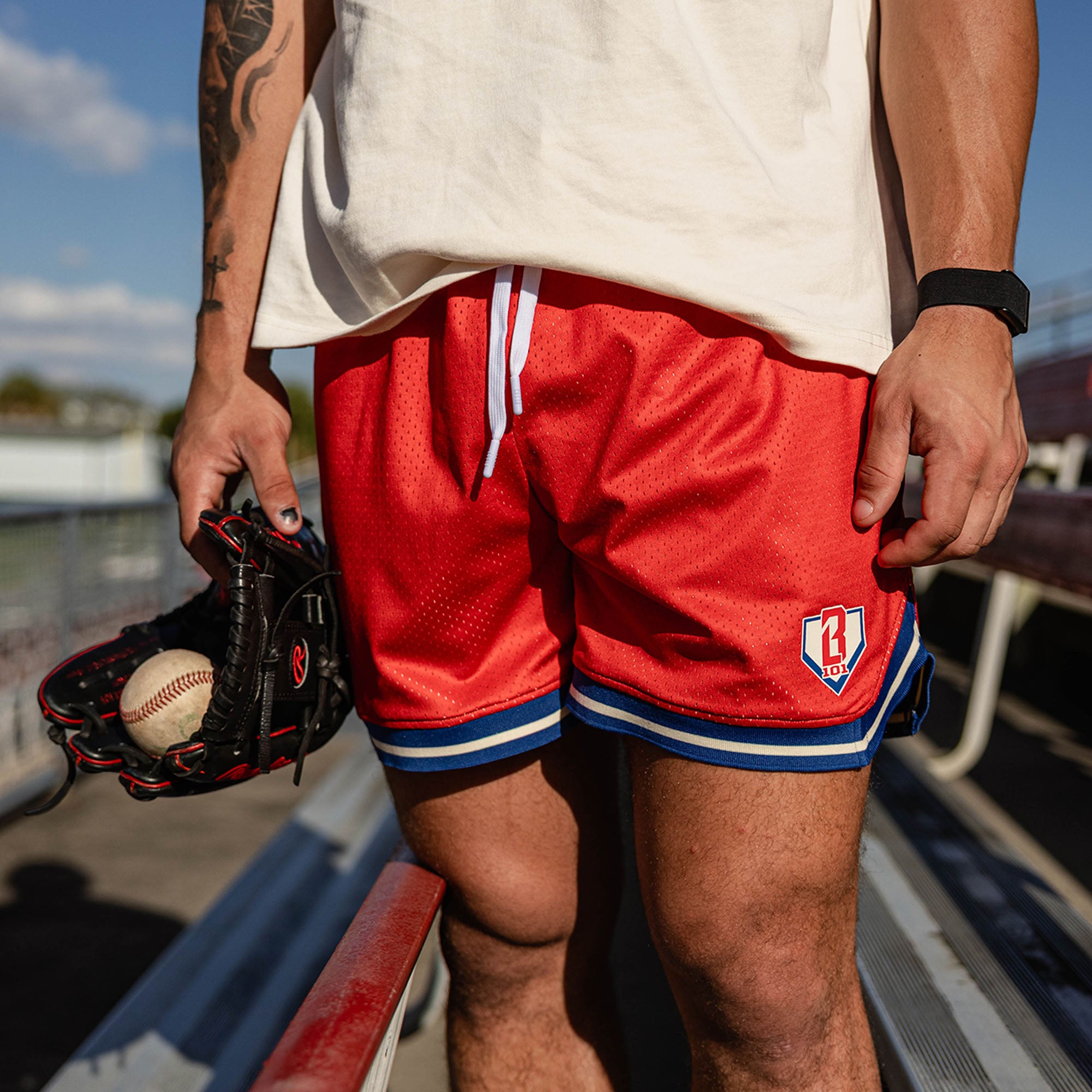 Close up of red mesh shorts with BL101 logo patch and blue and white striped hem paired with a cream tee with a glove and baseball held at the side