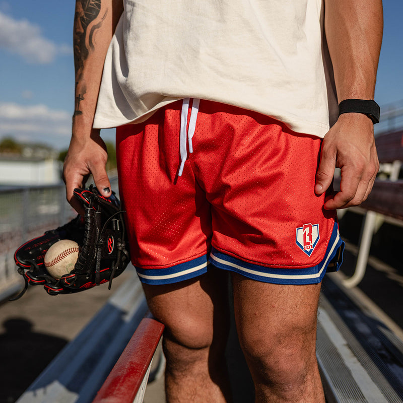 Close up of red mesh shorts with BL101 logo patch and blue and white striped hem paired with a cream tee with a glove and baseball held at the side