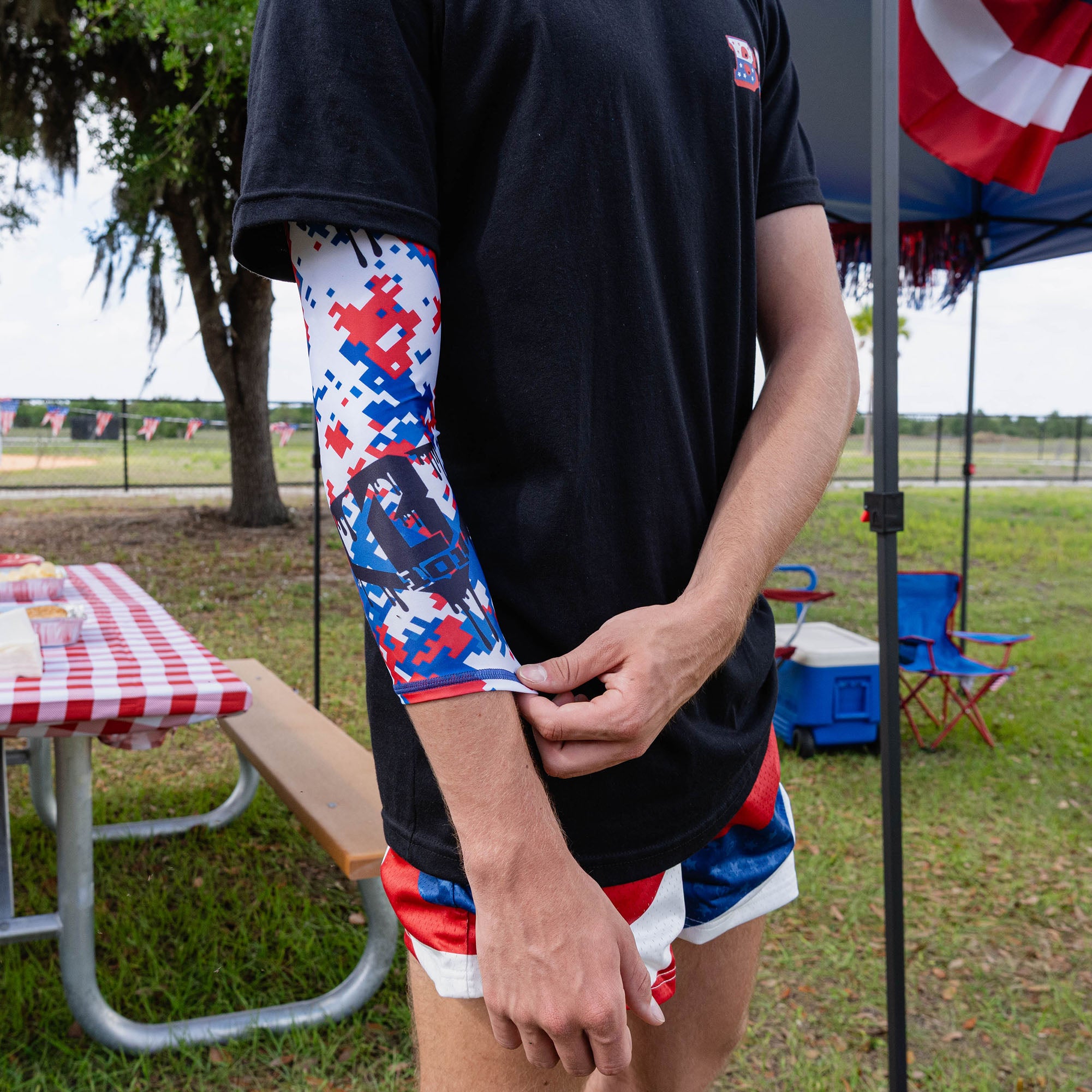 Adult male model adjusting wrist edge of red white and blue digital camo arm sleeve with black tee at outdoor baseball event