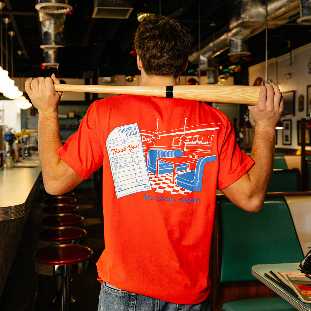 Person wearing a red t-shirt with a graphic design on the back featuring a restaurant scene and checkered floor.
