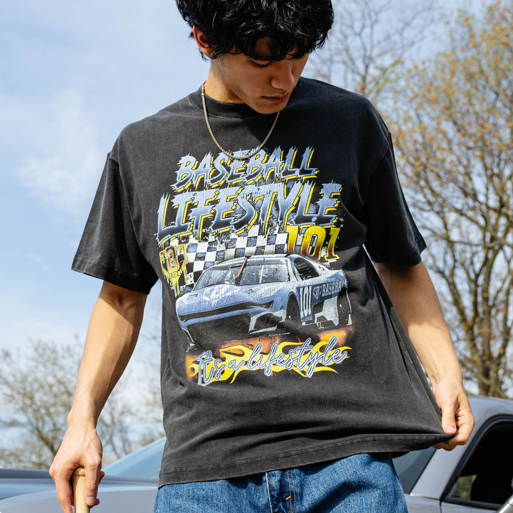 Close-up of washed black tee with vintage race car, checkered flag, and "It's a Lifestyle" flame graphic. Model holding baseball bat beside silver car.
