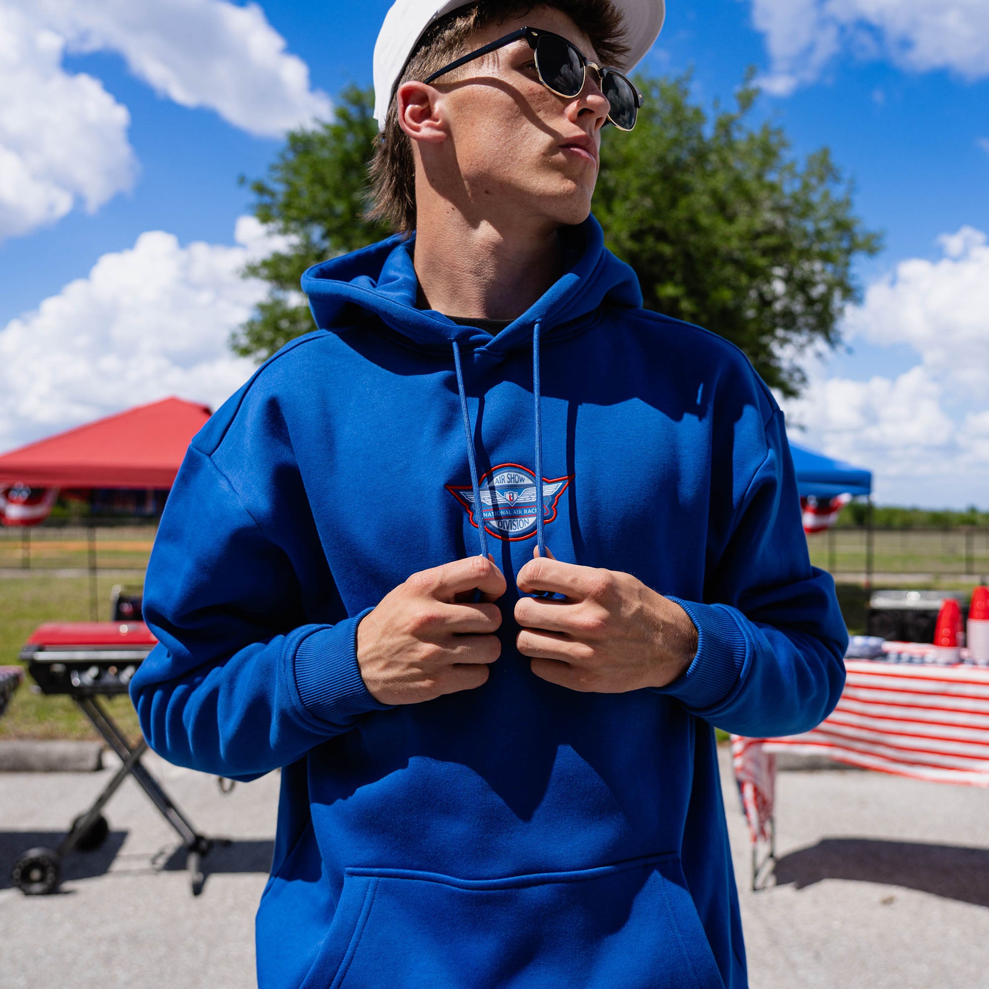 Close-up of adult male model in royal blue Fastball Flight hoodie pulling drawstrings showing Air Show Division badge on chest at outdoor event