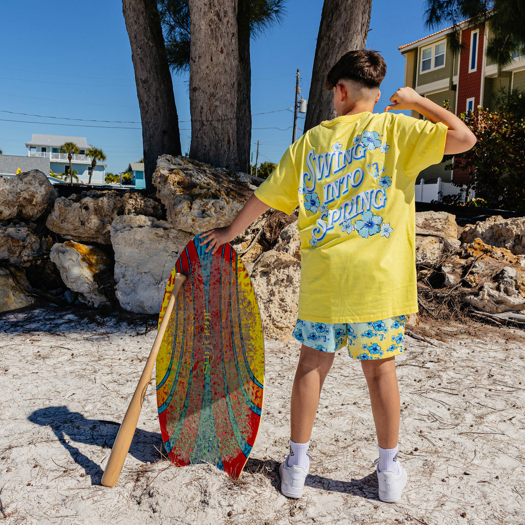 Person in yellow shirt with 'Swing into Spring' text and floral shorts holding a colorful board on a beach.