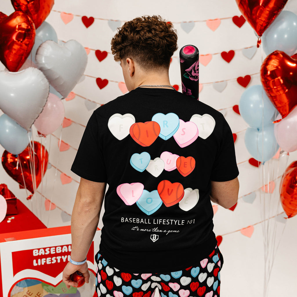 Person wearing a black t-shirt with colorful heart designs and heart-patterned pants, standing in front of heart-shaped balloons.
