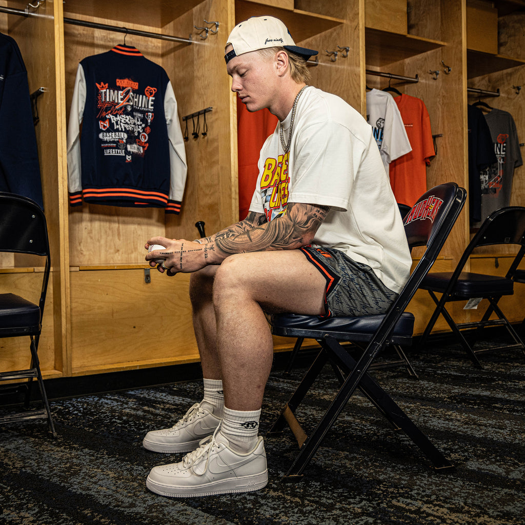 Person sitting in a locker room wearing a white t-shirt and shorts, surrounded by sports gear.