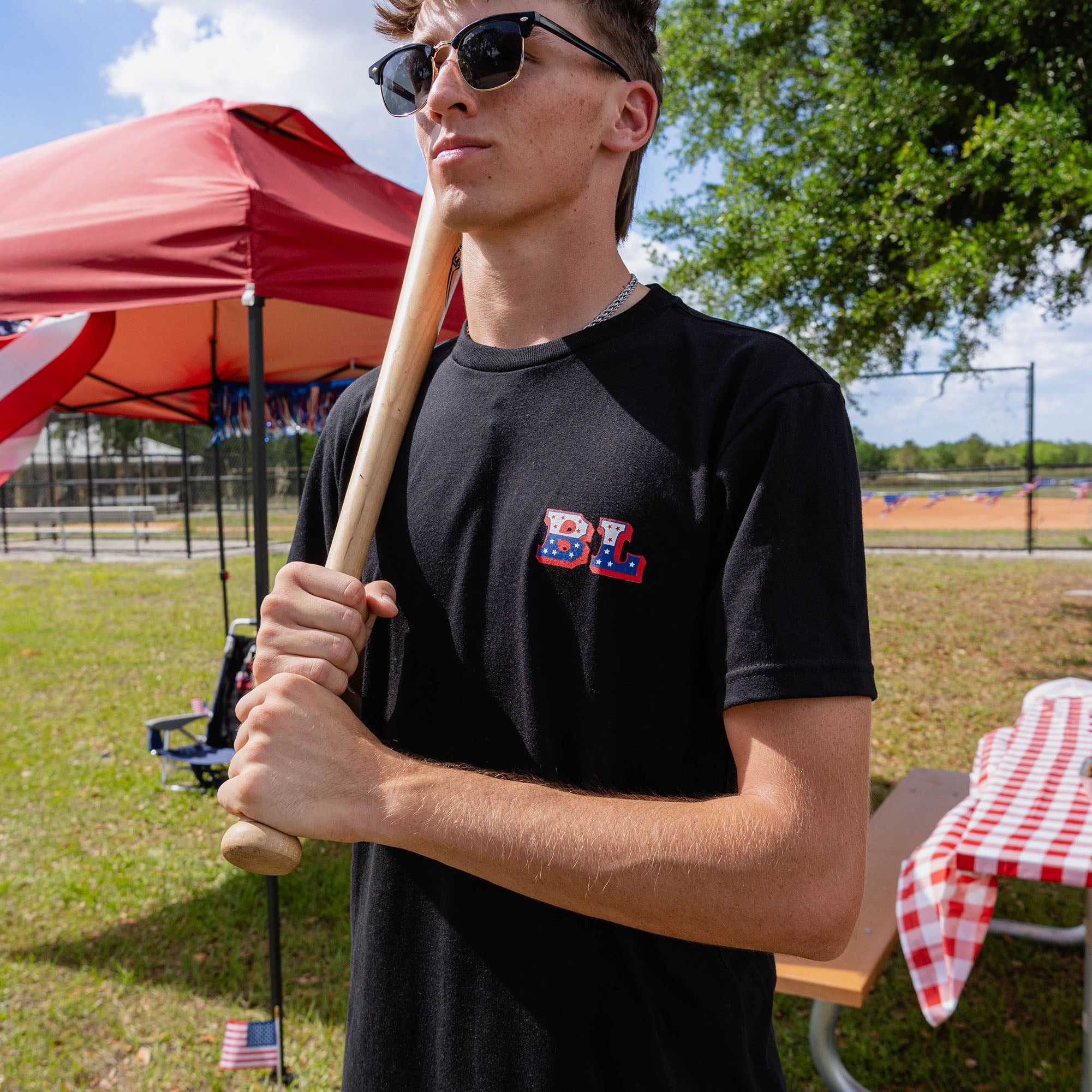 Close-up of adult male model wearing black Patriot Pitch tee holding baseball bat at shoulder at outdoor baseball event