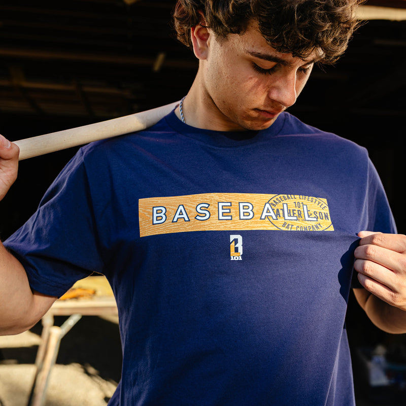 Close-up of a young adult male model pulling the front of the navy Radar Tee to show the wood grain baseball graphic and Father & Son Bat Company stamp design.