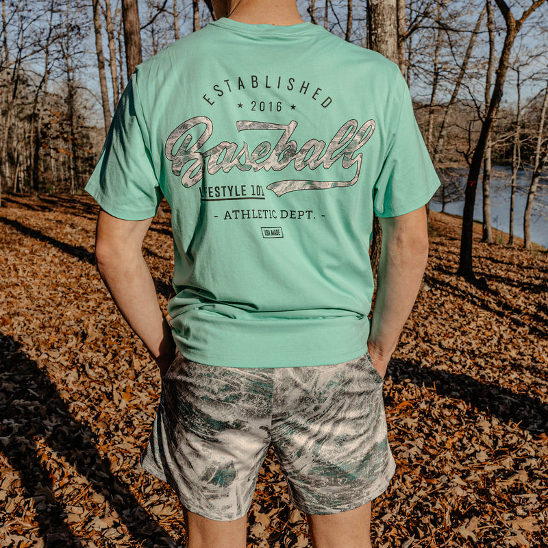 Person wearing a light green t-shirt with a 'Established 2016 Baseball Lifestyle 101' text in an outdoor setting with trees and a river.