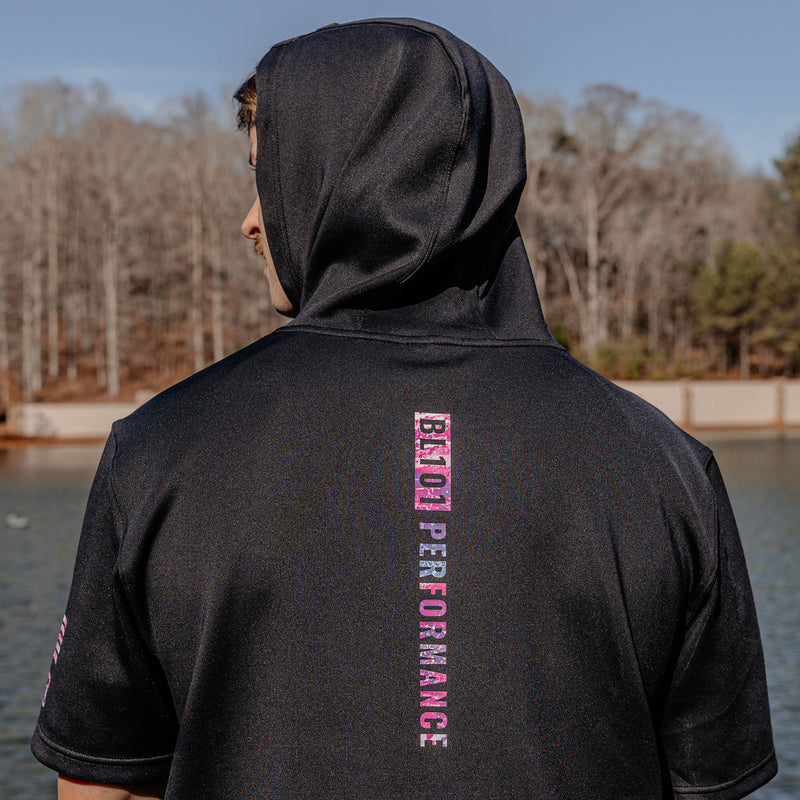 Person wearing a black hoodie with 'BL101 PERFORMANCE' text on the back, standing by a body of water.