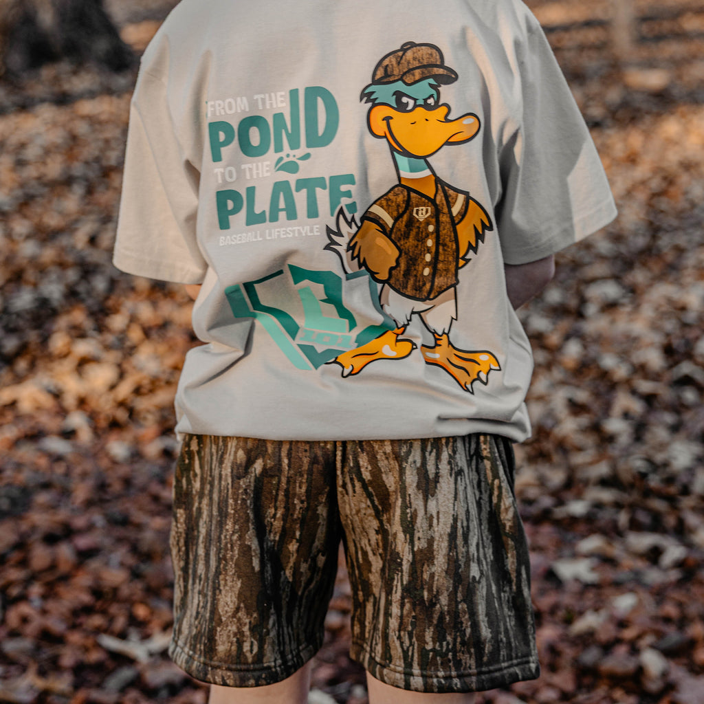 Person wearing a t-shirt with a cartoon duck design and 'From Pond to the Plate Baseball Lifestyle' text in a forest setting