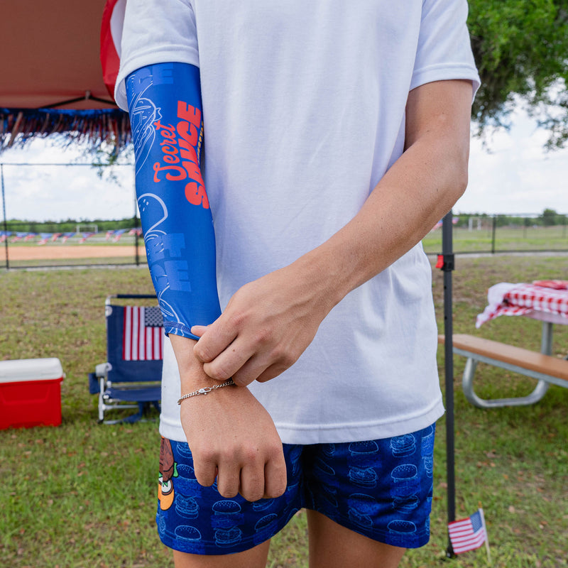 Adult male model adjusting wrist of blue Secret Sauce arm sleeve showing Secret Sauce logo detail while wearing white tee and blue Secret Sauce shorts at outdoor baseball event