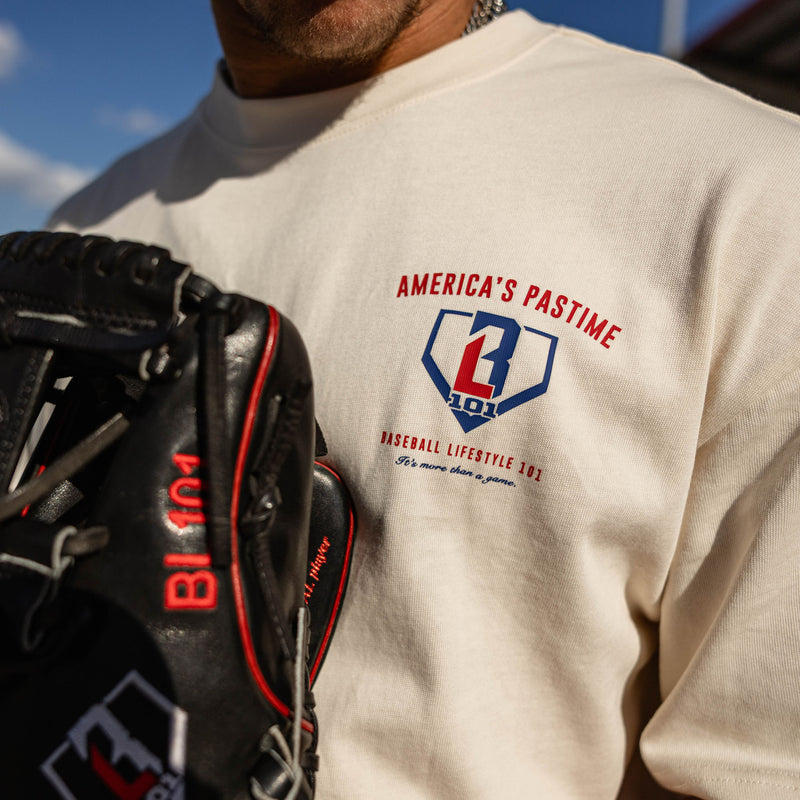 Close-up of the BL101 America's Pastime chest logo on the cream tee, with a baseball glove in the foreground.
