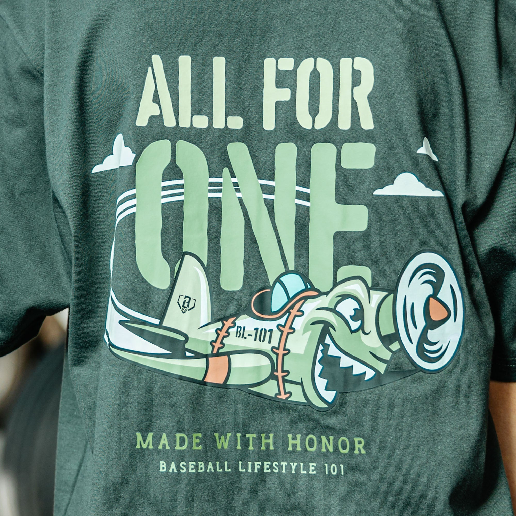 Close up of Dark Grey T- shirt with green cartoon airplane graphic and lettering that says 'ALL FOR ONE Made with honor Baseball Lifestyle 101' on person