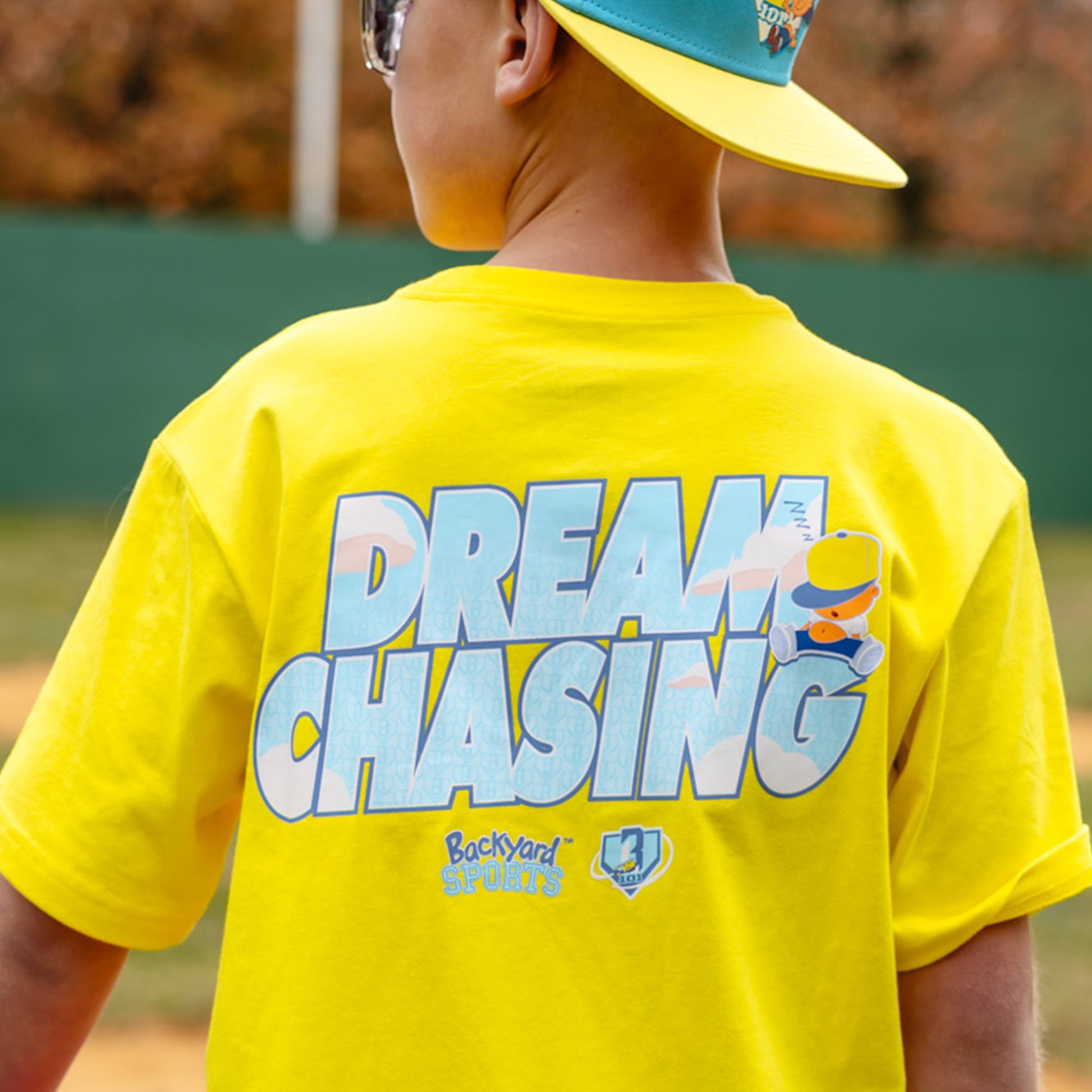 Person wearing a yellow 'Dream Chasing' t-shirt with a blue cap outdoors.