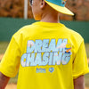 Person wearing a yellow 'Dream Chasing' t-shirt with a blue cap outdoors.