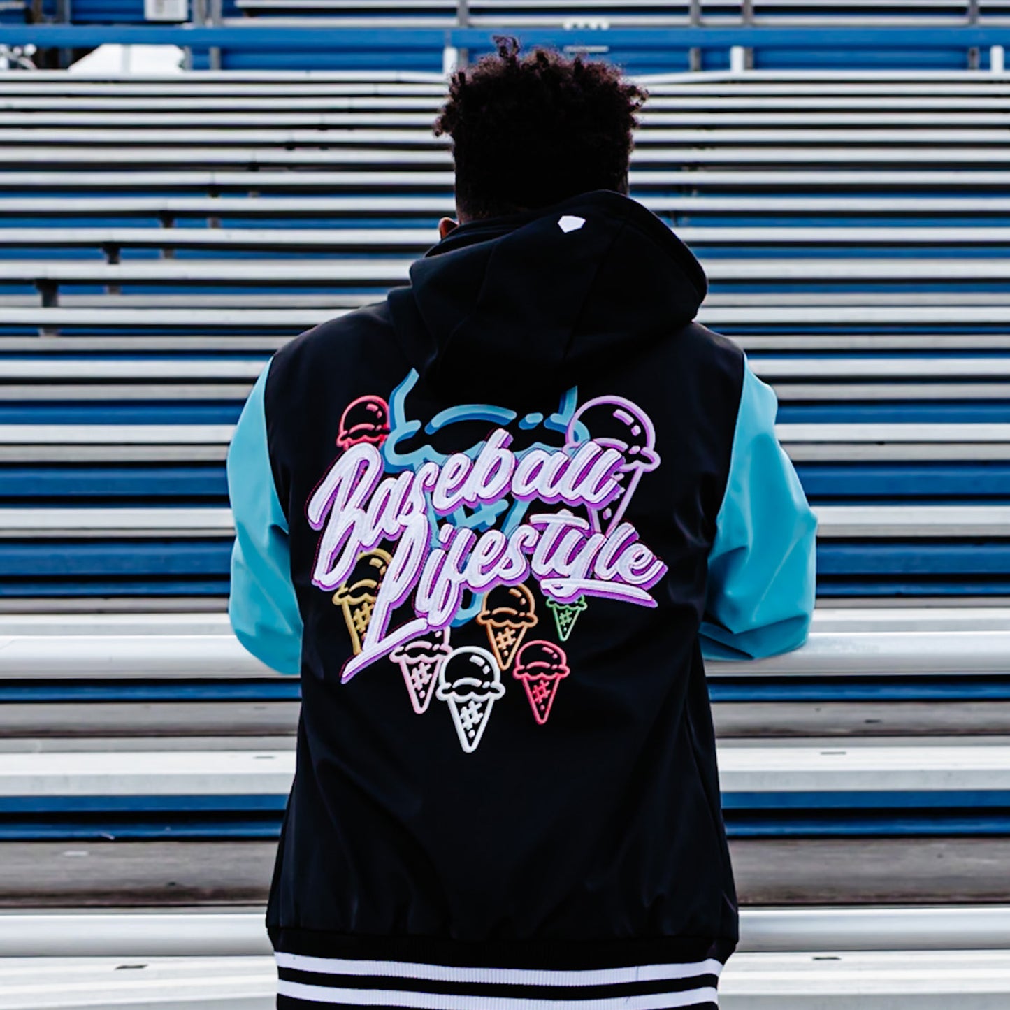 Person wearing a black and blue bomber jacket with 'Baseball Lifestyle' logo and colorful cones in front of bleachers.
