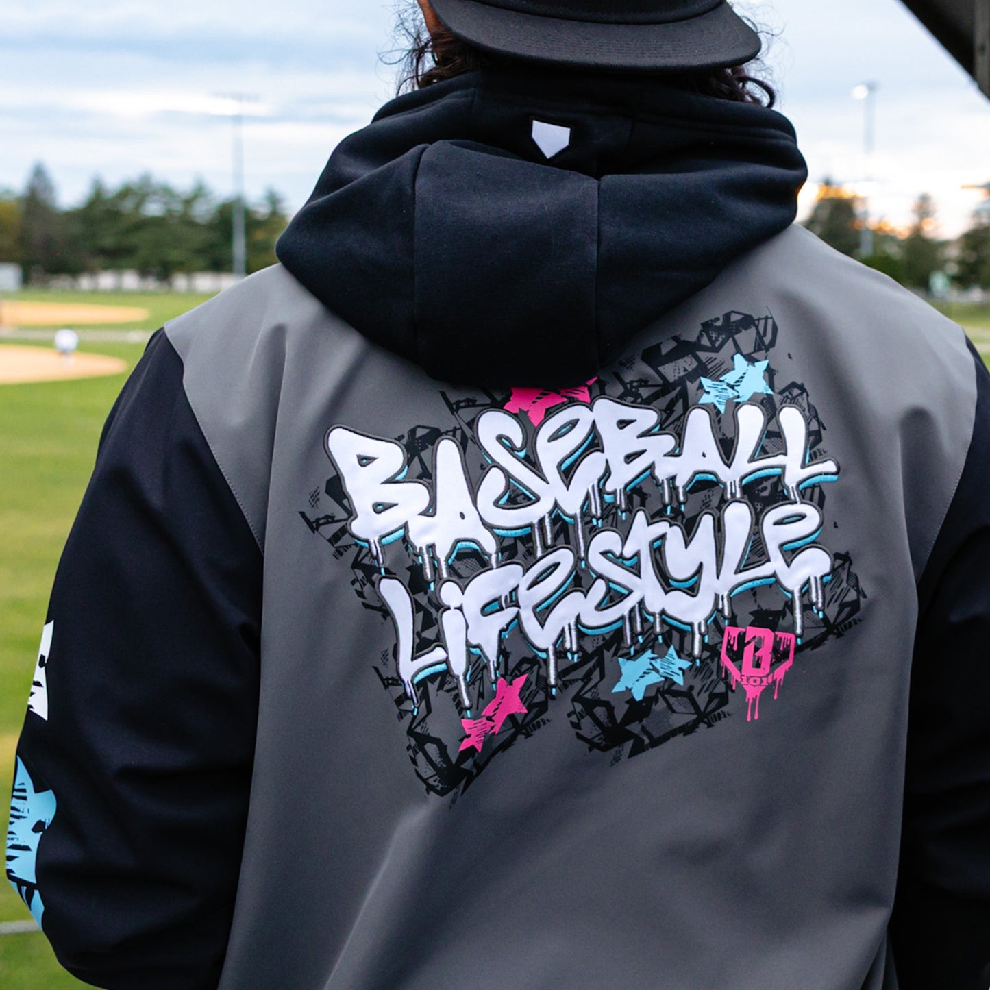 Person wearing a gray and black jacket with 'Baseball Lifestyle' design on the back, standing on a baseball field.