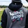 Person wearing a gray and black jacket with 'Baseball Lifestyle' design on the back, standing on a baseball field.