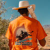 Person wearing an orange t-shirt with a bull graphic design and 'WHERE DUST MEETS DIAMOND' text, standing in a desert landscape.