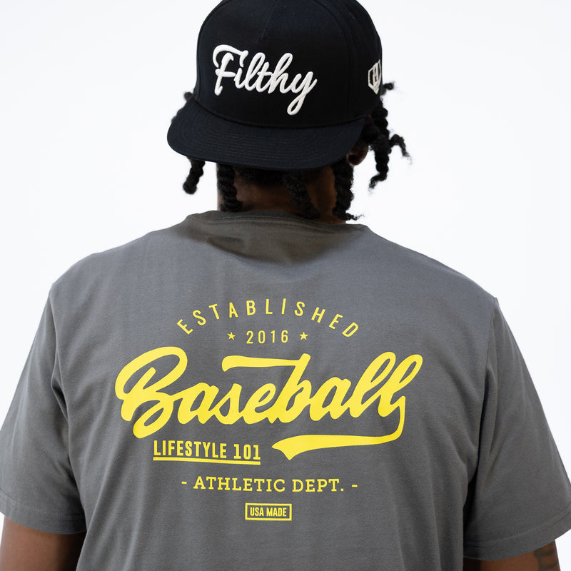 Person wearing a gray t-shirt with yellow 
'Baseball Lifestyle 101' text and a black cap with white 'Filthy' text 