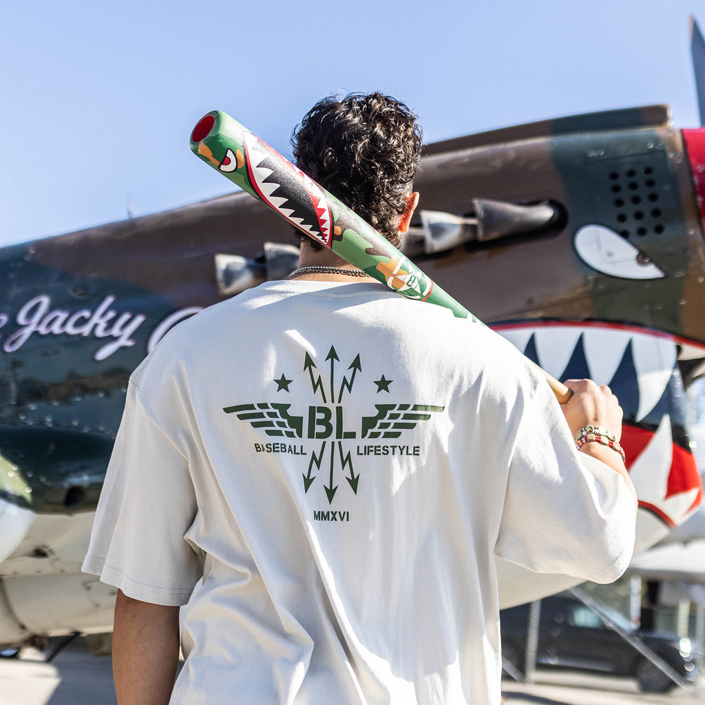 Person holding a baseball bat with a shark design, wearing a Tan t-shirt with dark green graphic featuring wings, arrows, and stars, with the Baseball Lifestyle BL logo in the center