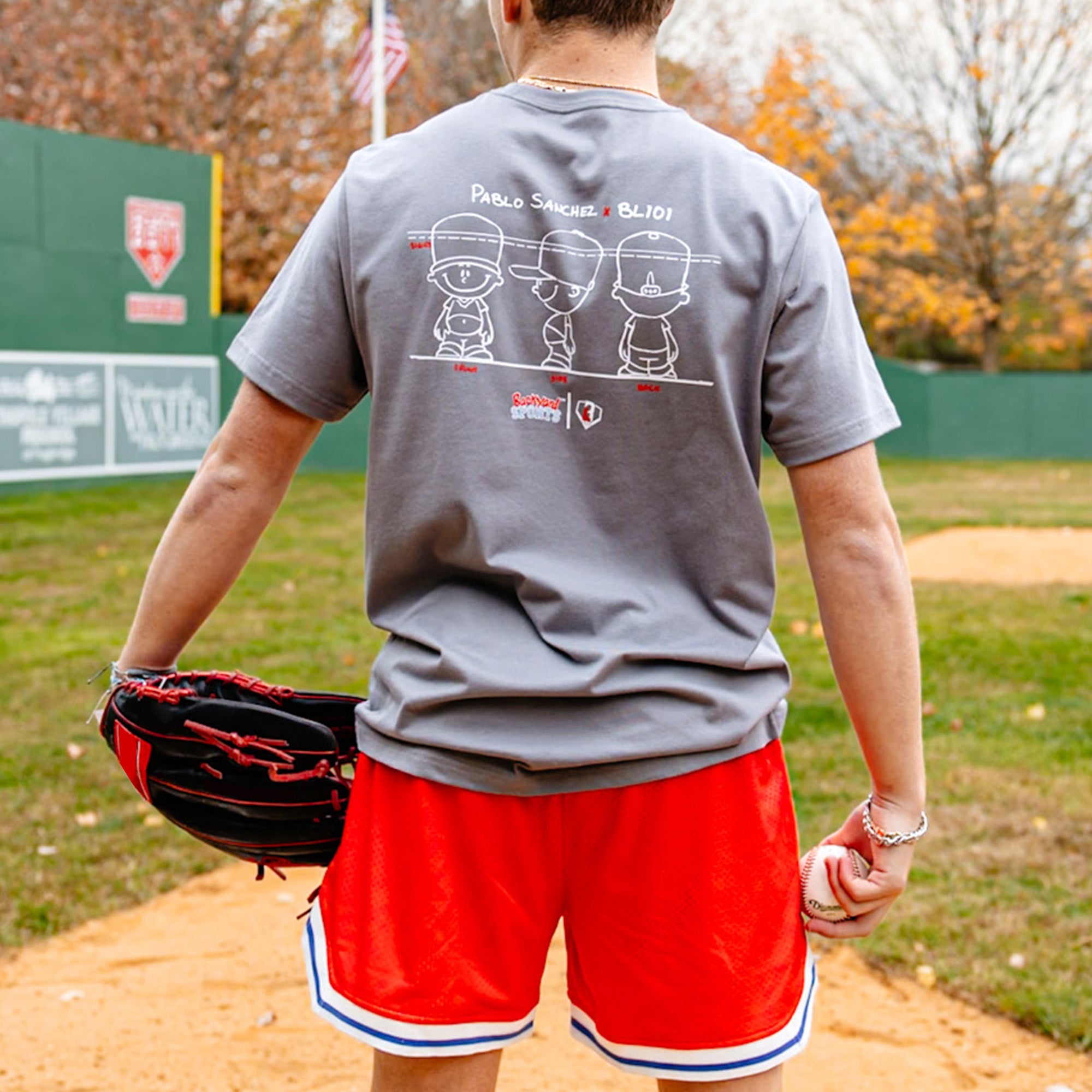 Person on a baseball field wearing a gray t-shirt with Pablo Sanchez graphics and red shorts, holding a baseball and glove.