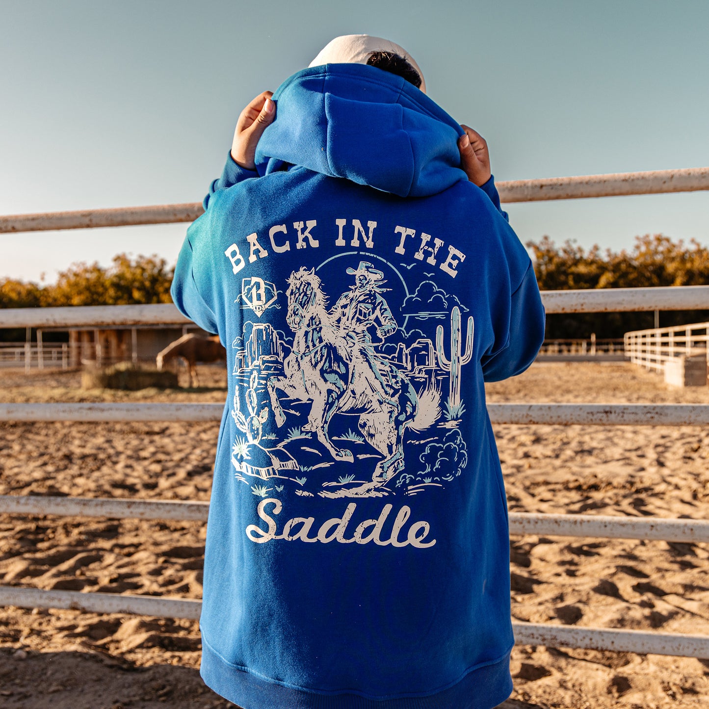 Person wearing a blue hoodie with 'Back in the Saddle' text and graphic, standing in an outdoor setting.