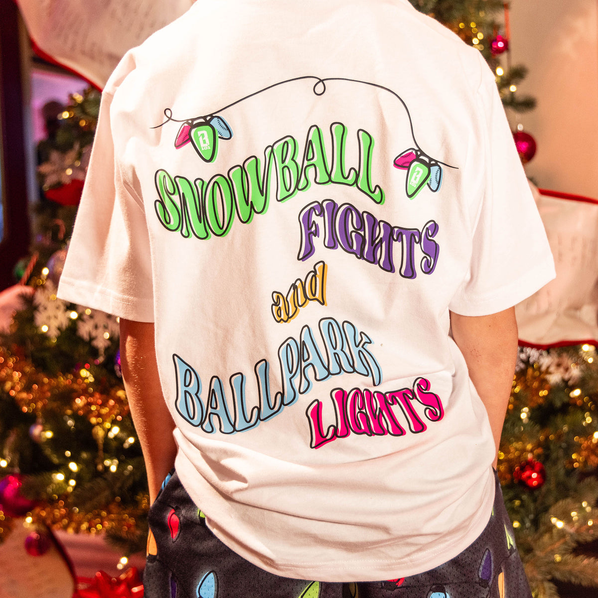 Person wearing white t-shirt with colorful 'Snowball Fights and Ballpark Lights' text design