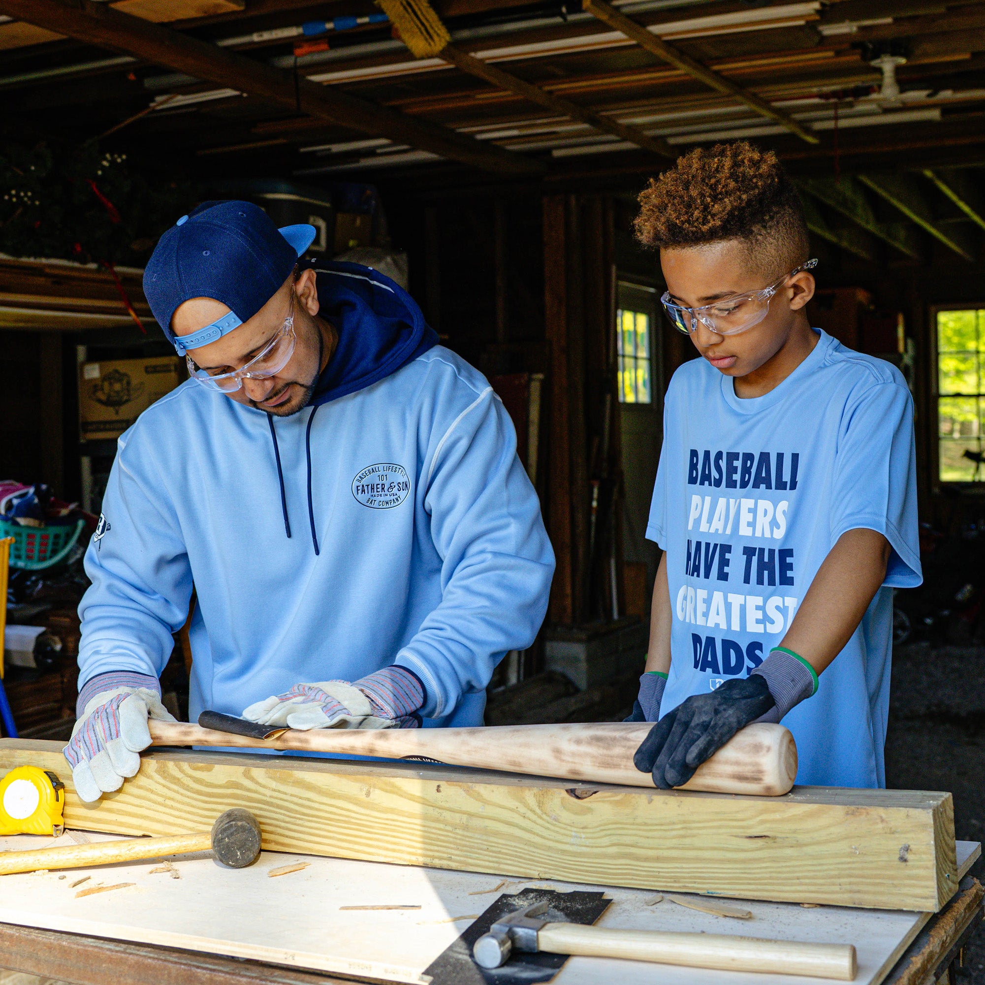 Father and son working side by side at a woodshop bench crafting a baseball bat while wearing Father & Son Bat Company apparel including the navy snapback worn backwards.