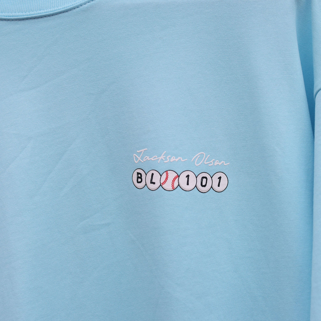Close-up of the chest graphic on the light blue BSBL Era tee, showing the "Jackson Olson" script above the BL101 baseball bead logo