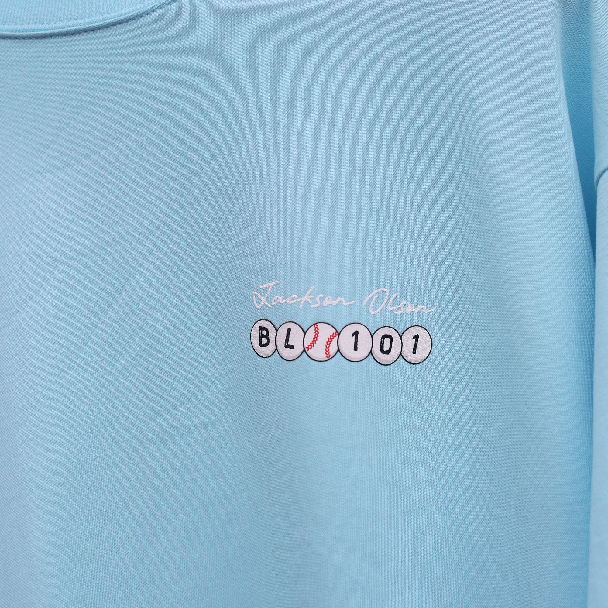 Close-up of the chest graphic on the light blue BSBL Era tee, showing the "Jackson Olson" script above the BL101 baseball bead logo