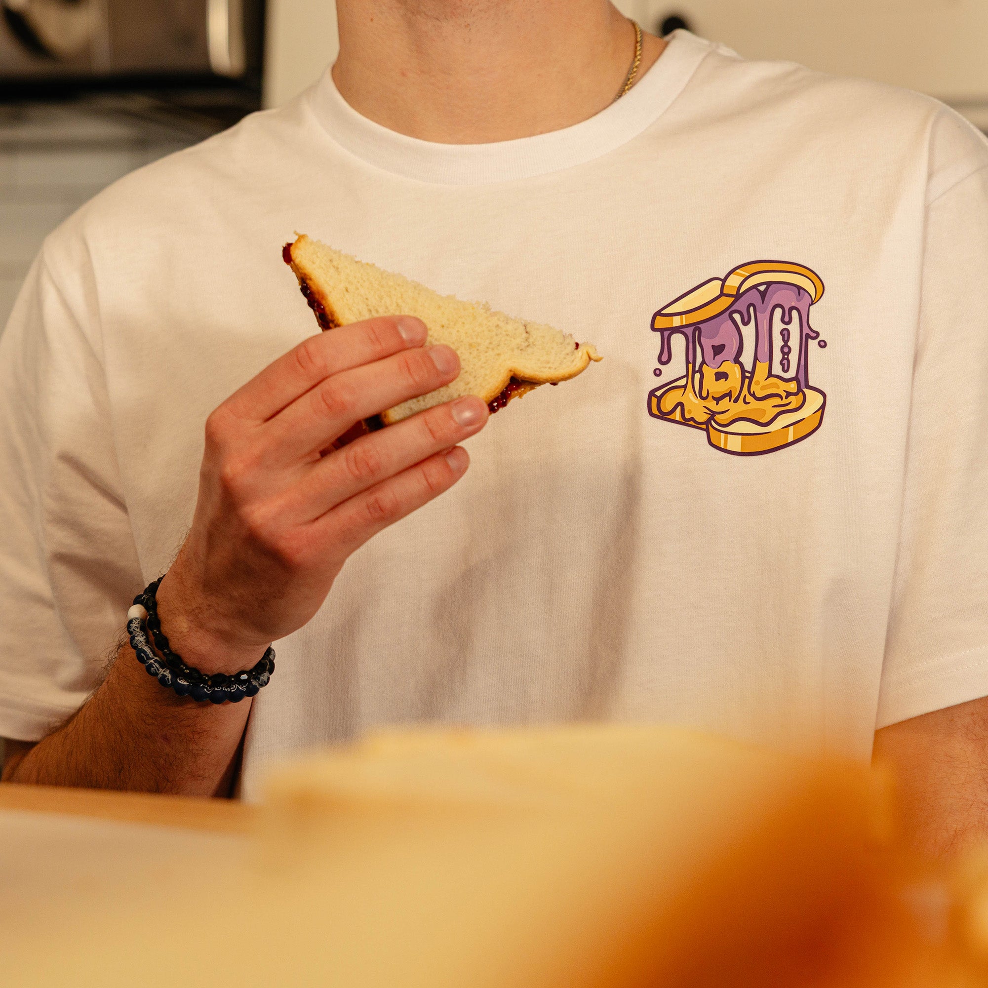 Close-up of the BL101 Peanut Butter and Jelly chest logo on the white tee, with a PB&J sandwich held in the foreground.