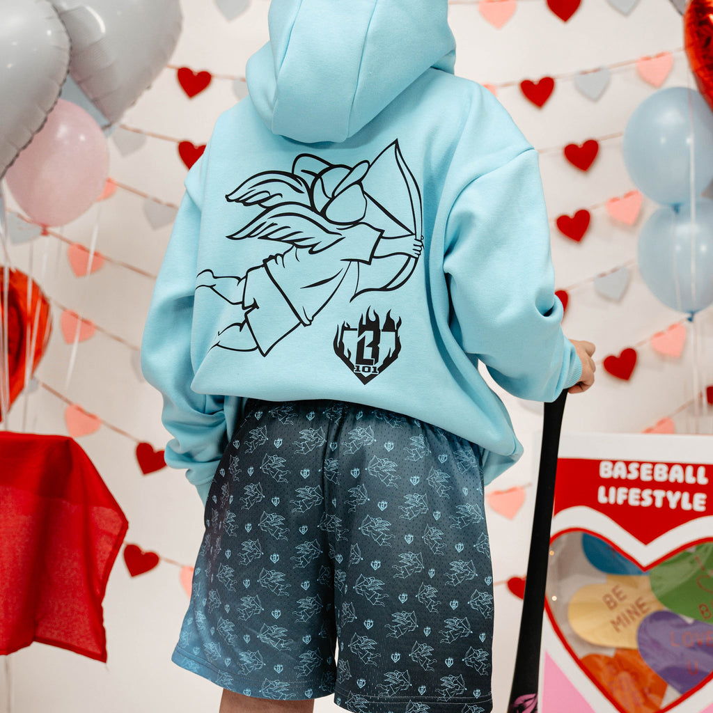 Person wearing a light blue hoodie with a boy holding arrow graphic design and gradient blue shorts, standing in front of a heart-themed wall with balloons.