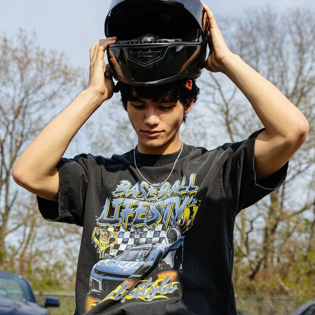 Young man putting on black racing helmet, wearing washed black Fast Track Fielder tee with full race car graphic, outdoors.