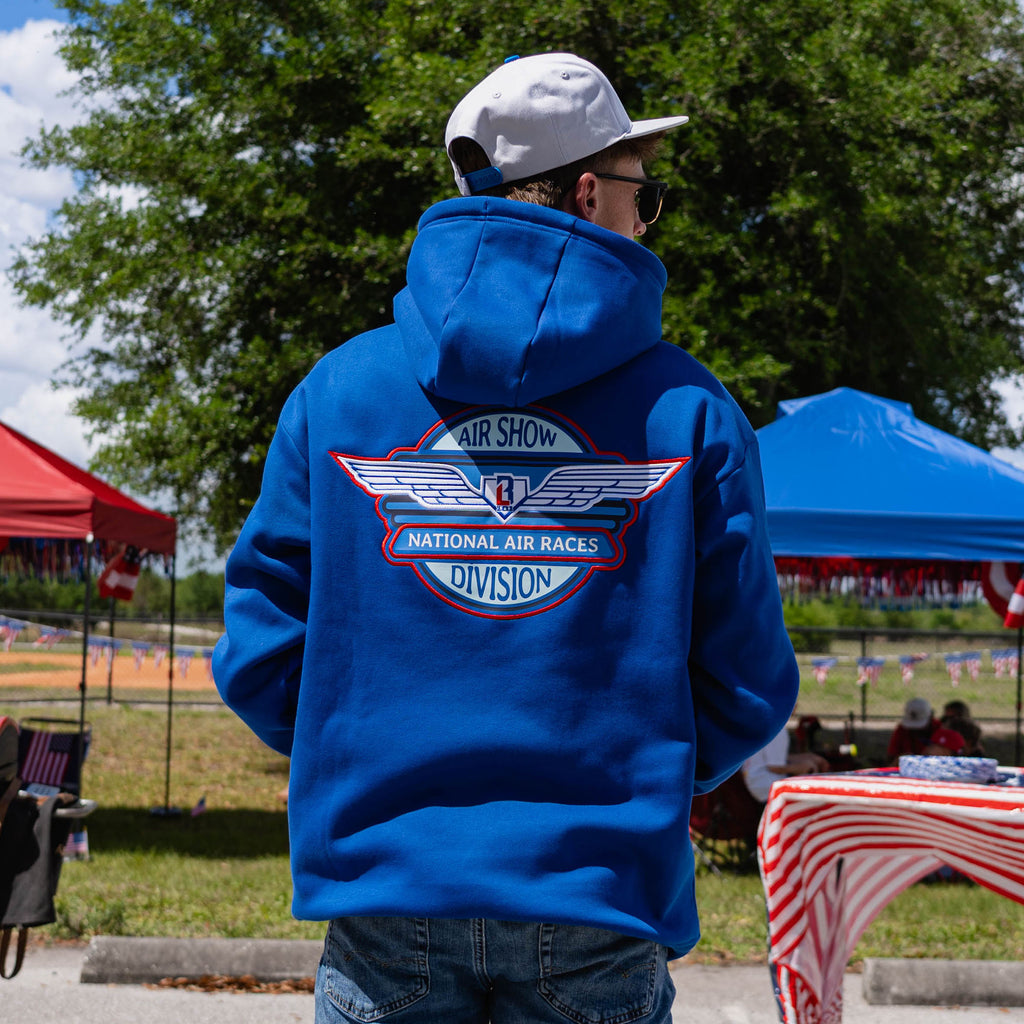 Back view of adult male model showing full Air Show National Air Races Division badge on royal blue hoodie at outdoor baseball event