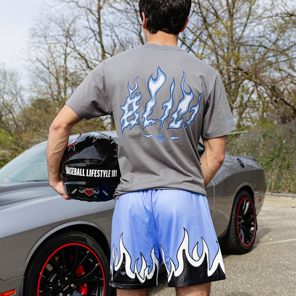 Back view of gray Flame Thrower tee and blue flame mesh shorts with white and black flame hem graphic, model holding Baseball Lifestyle 101 helmet beside gray Dodge Challenger.