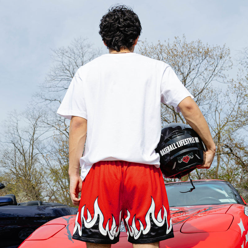 Back view of red flame mesh shorts with white and black flame hem graphic, model holding Baseball Lifestyle 101 helmet beside red Corvette.