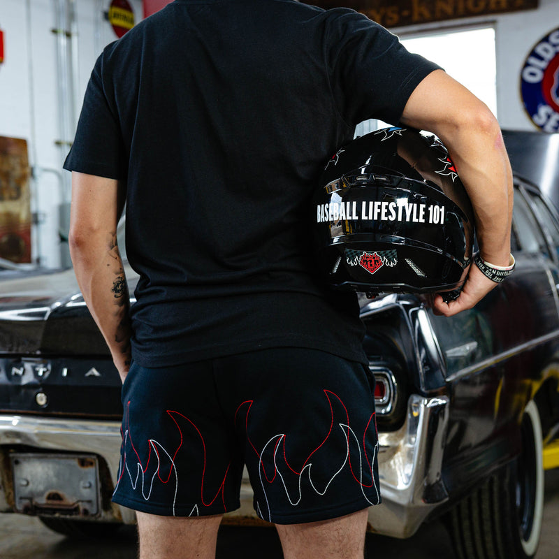 Back view of black flame sweatshorts showing red and white stitched flame hem graphic, model holding Baseball Lifestyle 101 helmet in vintage garage.