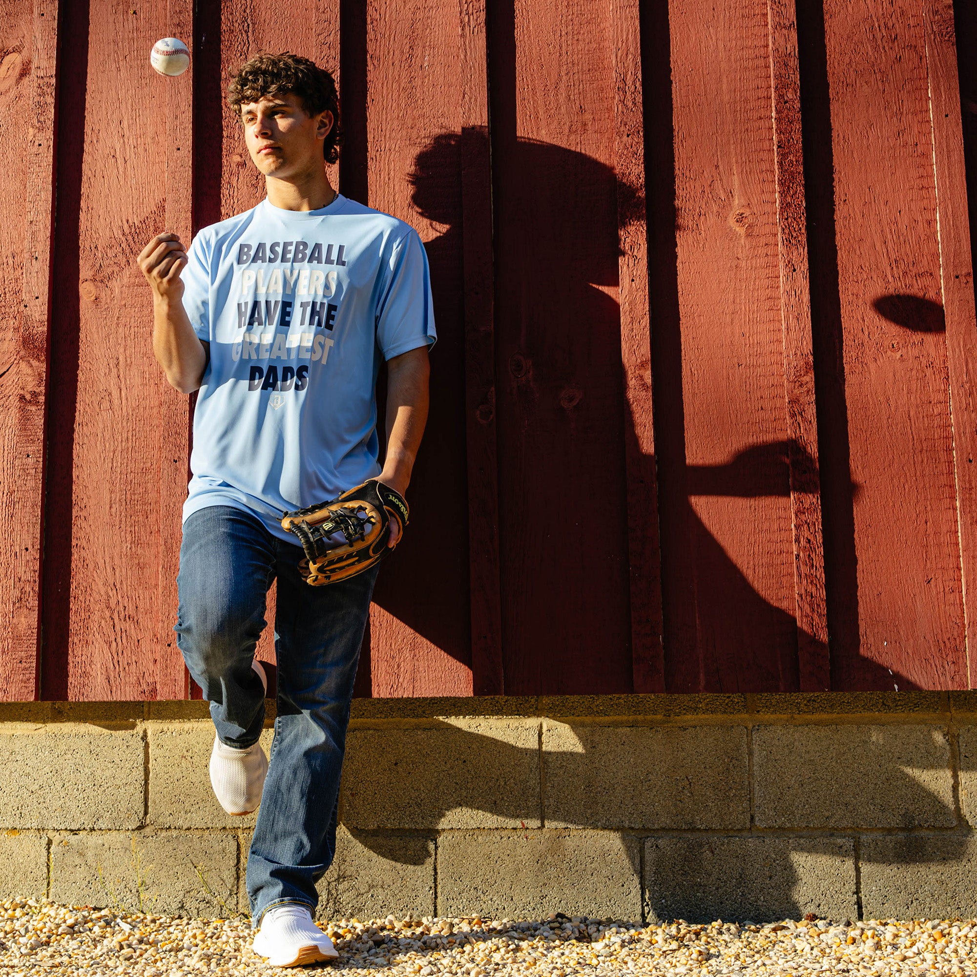 Young adult male model leaning against a red barn wall with a fielding glove and a baseball mid-air while wearing the columbia blue Greatest Dad Athletic Tee paired with jeans and white sneakers.