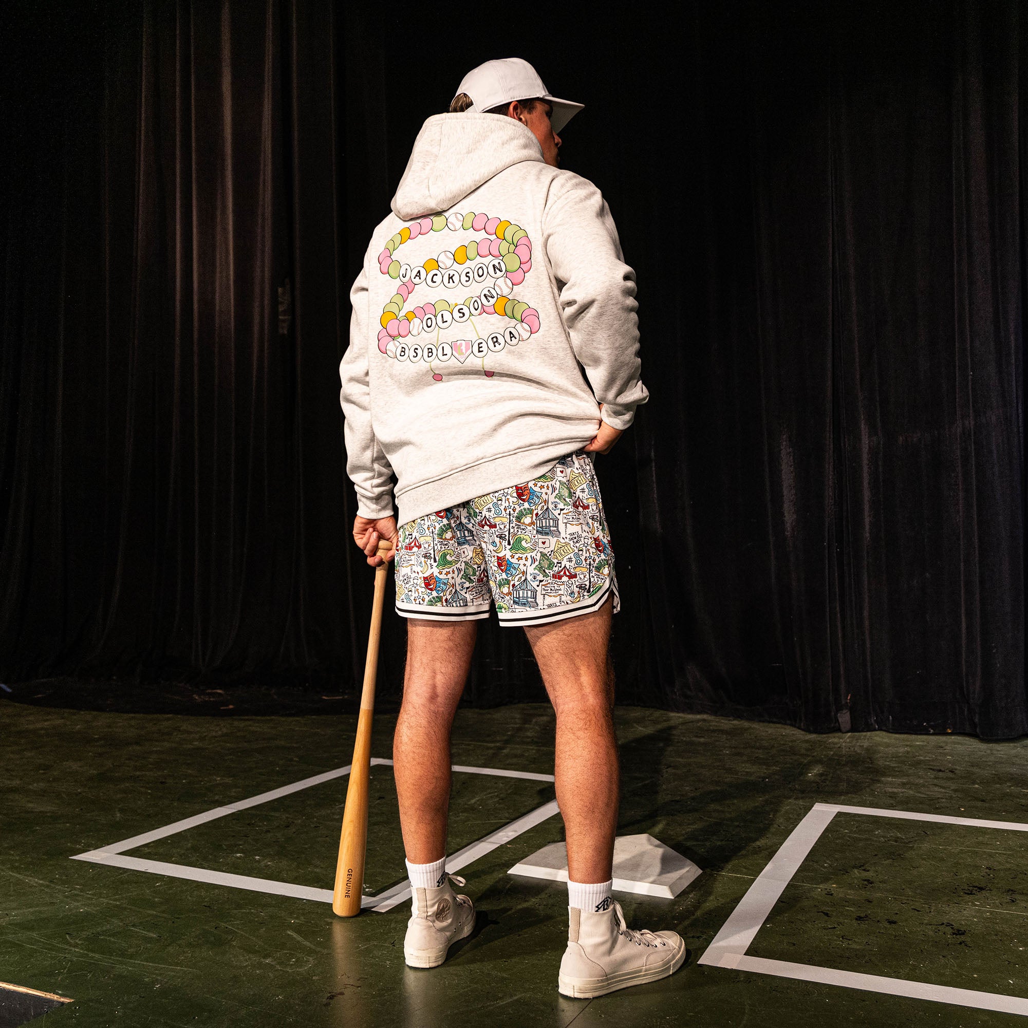 Back view of the adult male model wearing the white Jackson Olson x BL101 Hometown Shorts paired with the BSBL Era hoodie showing the friendship bracelet back graphic