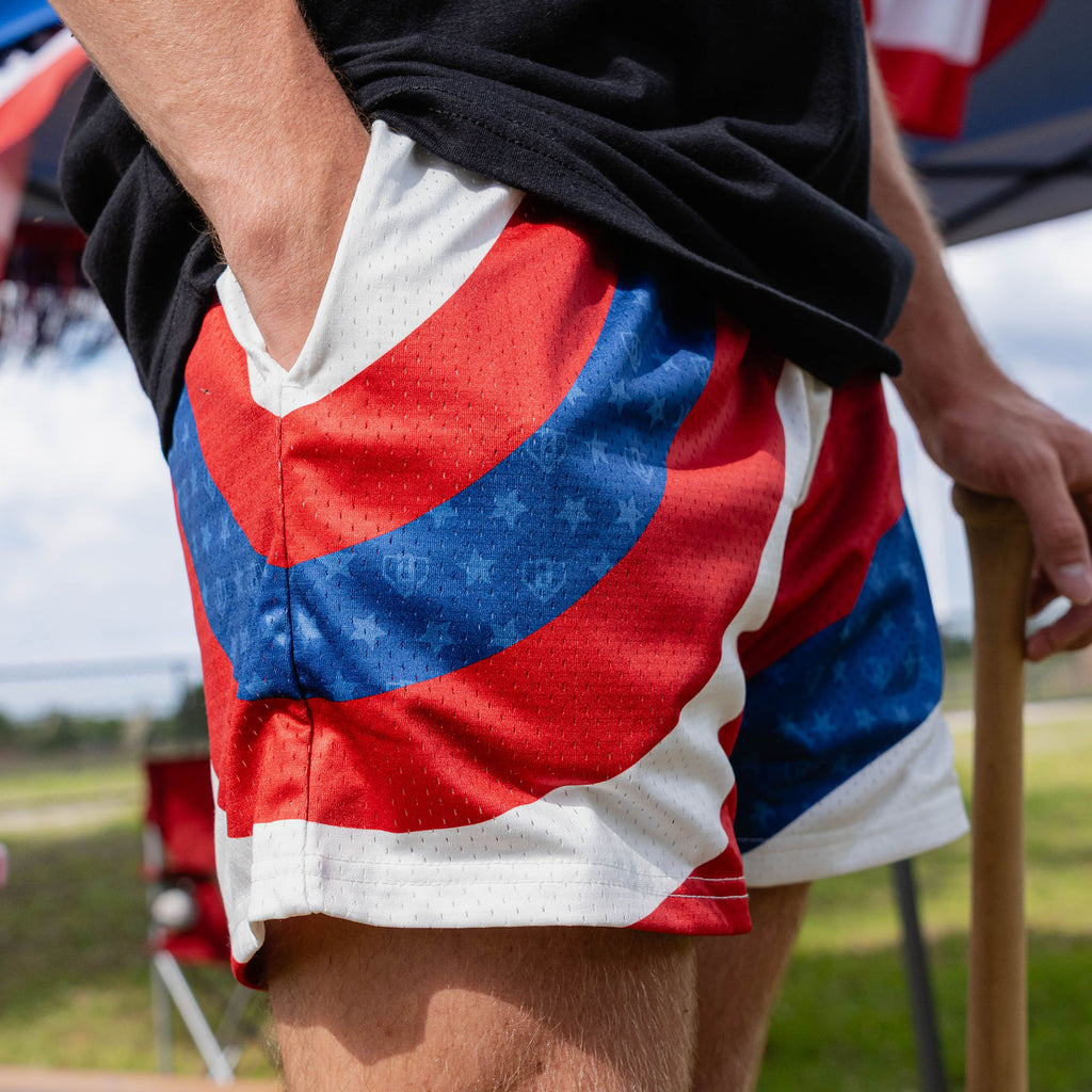 Lower body close-up of adult male model in red white and blue Patriot Pitch shorts showing diagonal colorblock detail with hand in pocket at outdoor event