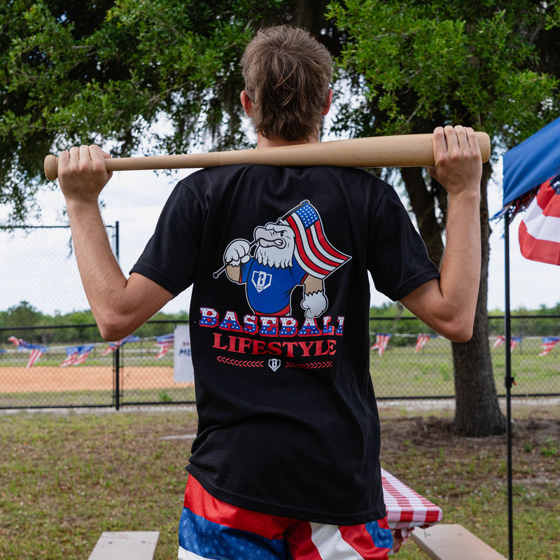 Back view of adult male model holding baseball bat across shoulders showing Baseball Lifestyle eagle graphic on black tee at outdoor baseball event