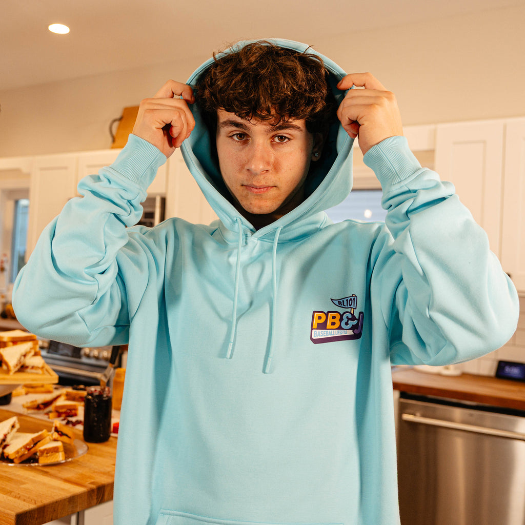 Male model pulling up the hood of the BL101 PB&J Baseball Lifestyle light blue hoodie in a kitchen, showing the chest logo.
